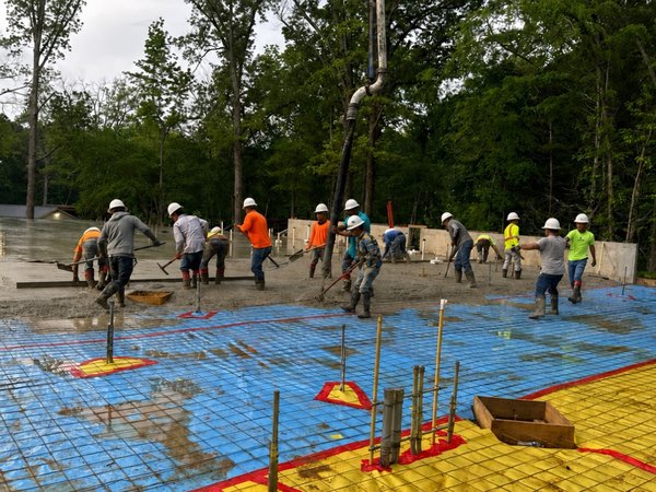 Crew hand-finishing a large concrete slab pour as a pump truck boom places fresh mix overhead