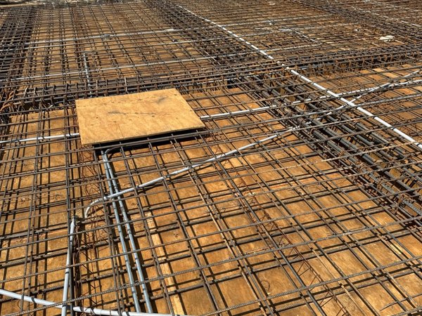 Wide view of rebar mesh placed for a large slab pour, with campus buildings and a ballfield visible beyond the site