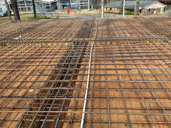 Rebar mesh placed over a thickened footing trench, with structural steel framing rising in the background