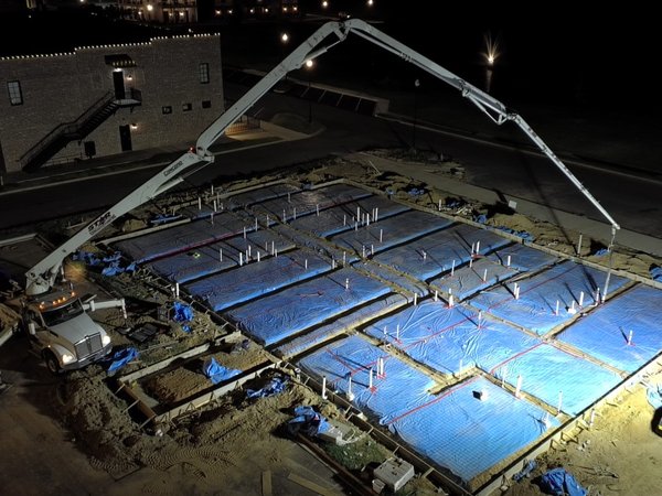 Nighttime aerial view of a concrete slab pour at Silo Square, with a pump truck boom extended over the site next to a brick building