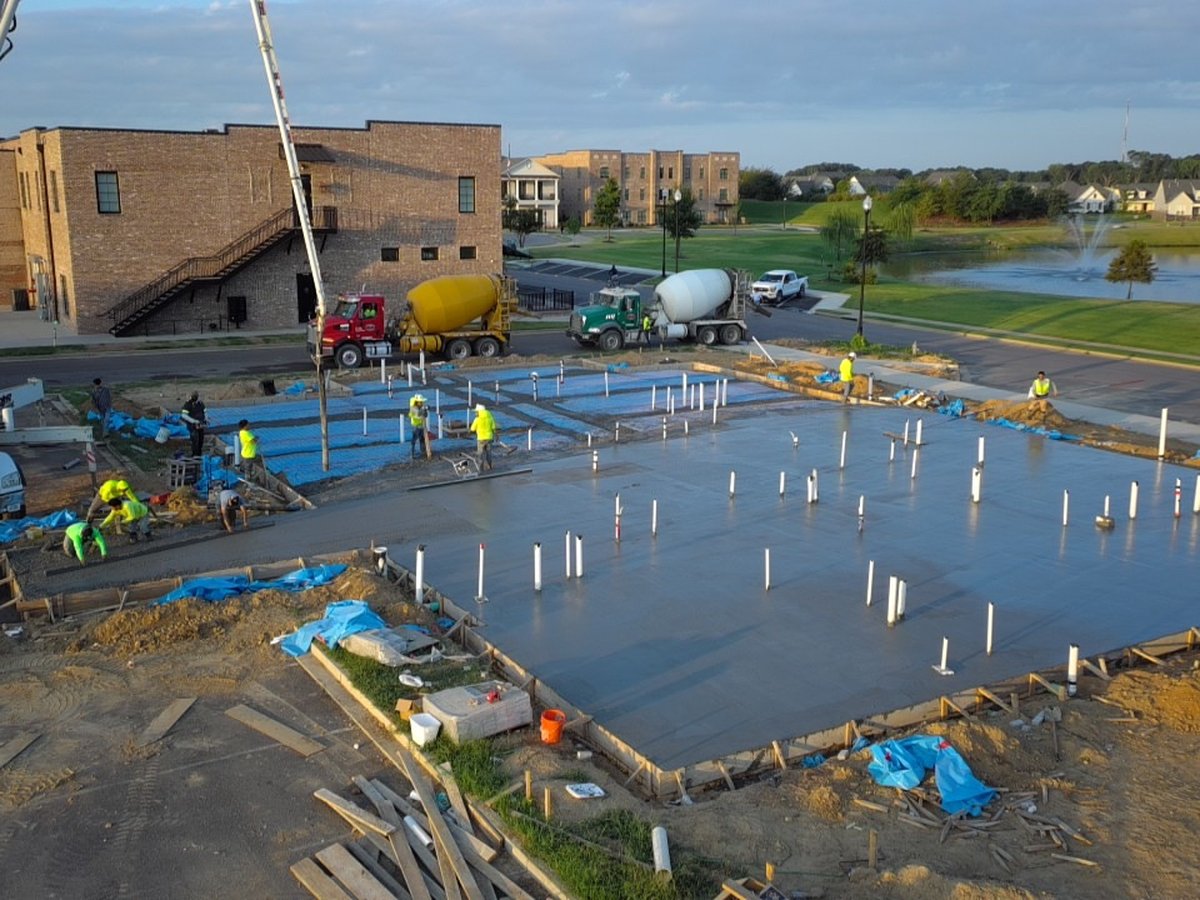 Daytime aerial view of a large concrete slab pour at Silo Square, with a pump truck and crew working beside completed brick buildings and a pond in the background